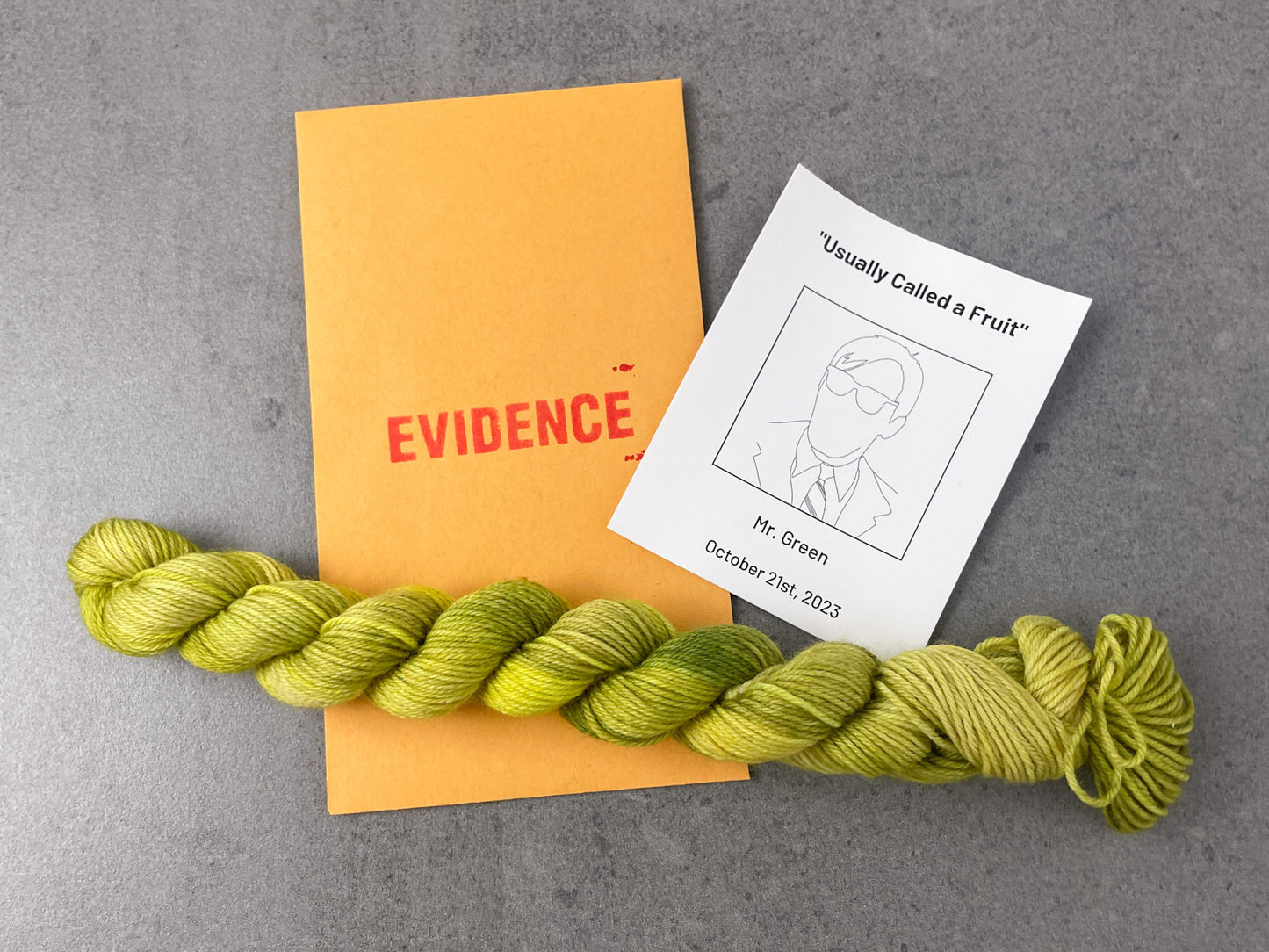 A tonal green skein of yarn on top of an envelope stamped with "Evidence" and a card with a drawing of Mr. Green on it.