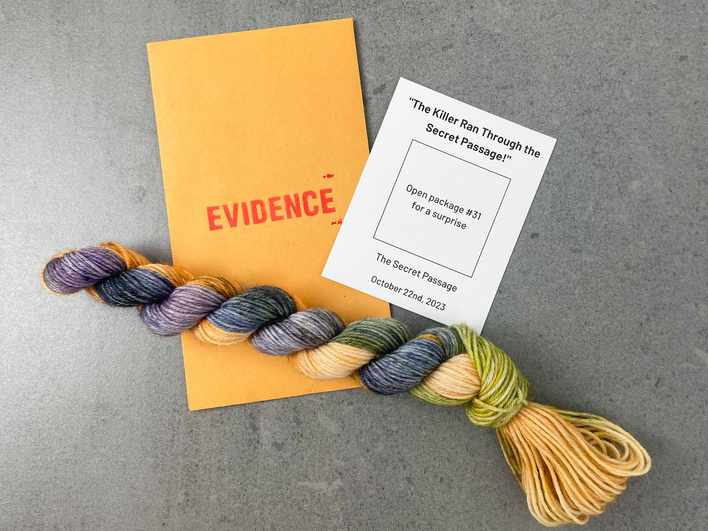 A variegated skein of purple, blue, green, and orange yarn on top of an envelope stamped with "Evidence" and a white card with colorway information on it.