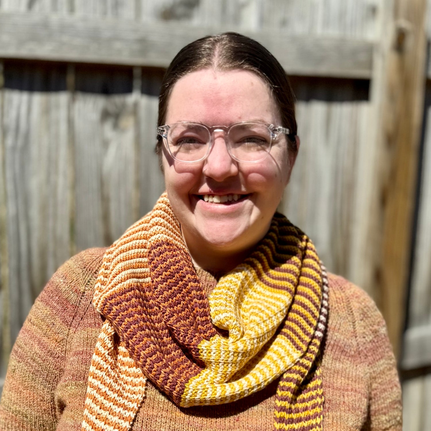 Cat, a white woman, smiles at the camera. She's wearing a handspun knit sweater in fall colors and a knit striped scarf with orange, yellow, red, and white yarn.