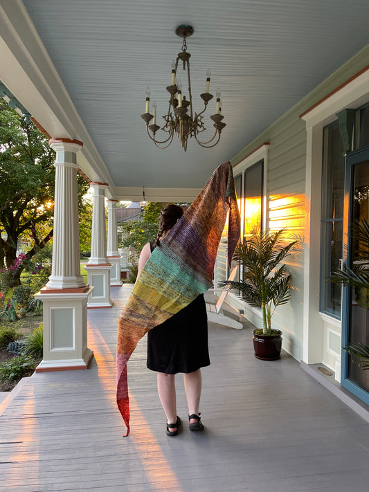 Cat holds up a rainbow shawl on a front porch with a large iron chandelier.