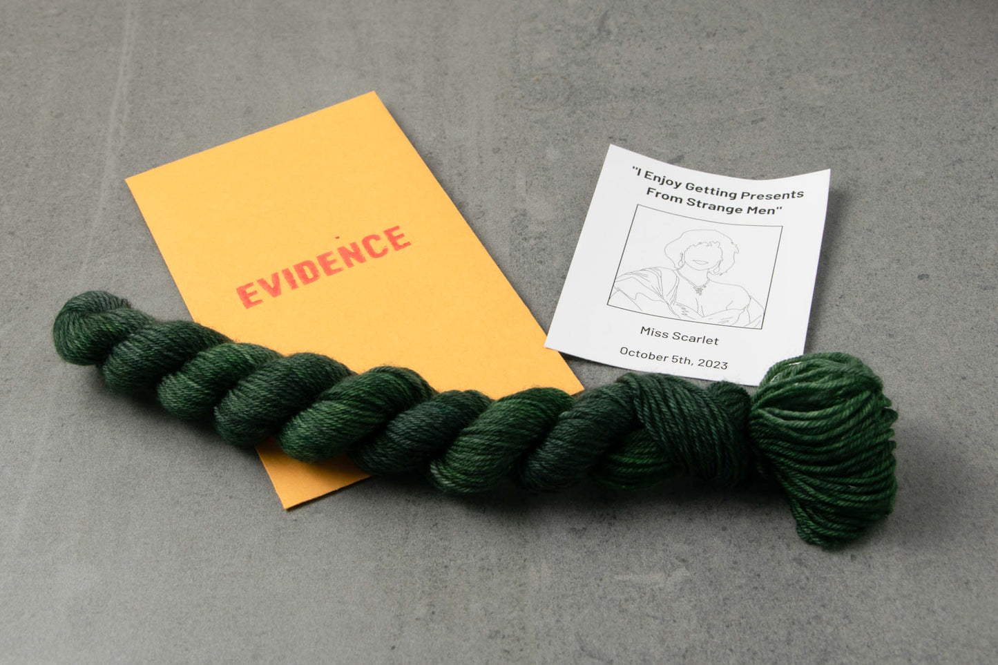 A skein of deep green on top of an envelope stamped with "Evidence" and a card with a drawing of Miss Scarlet on it.