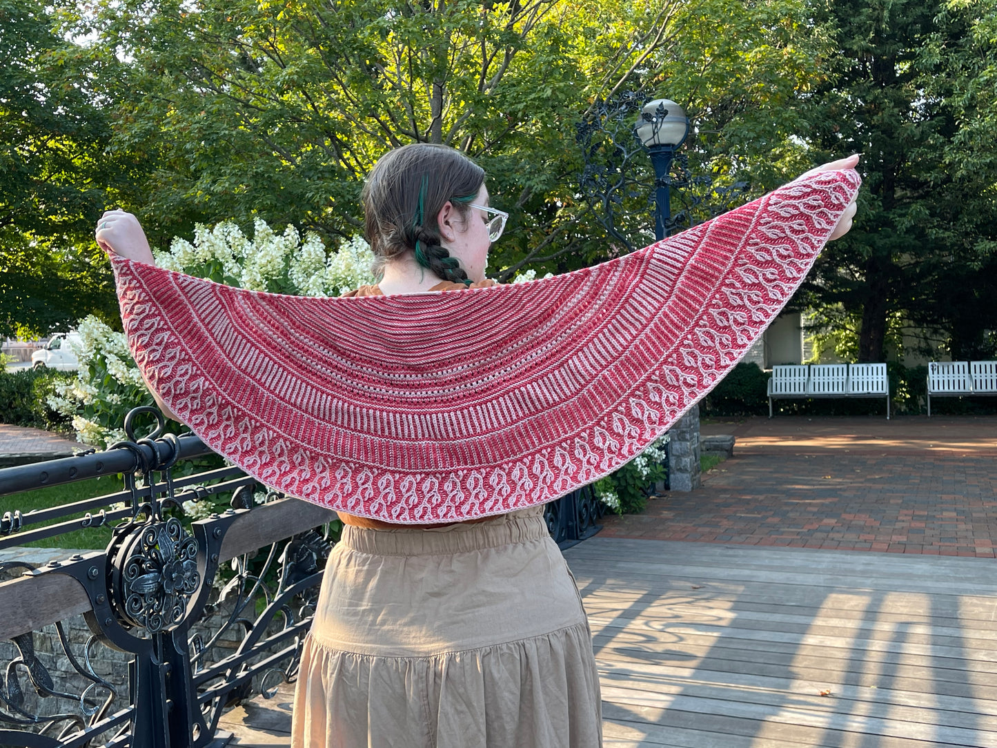 Cat, a white woman, stands facing away from the camera and holds out the Gradual Brioche shawl behind her.