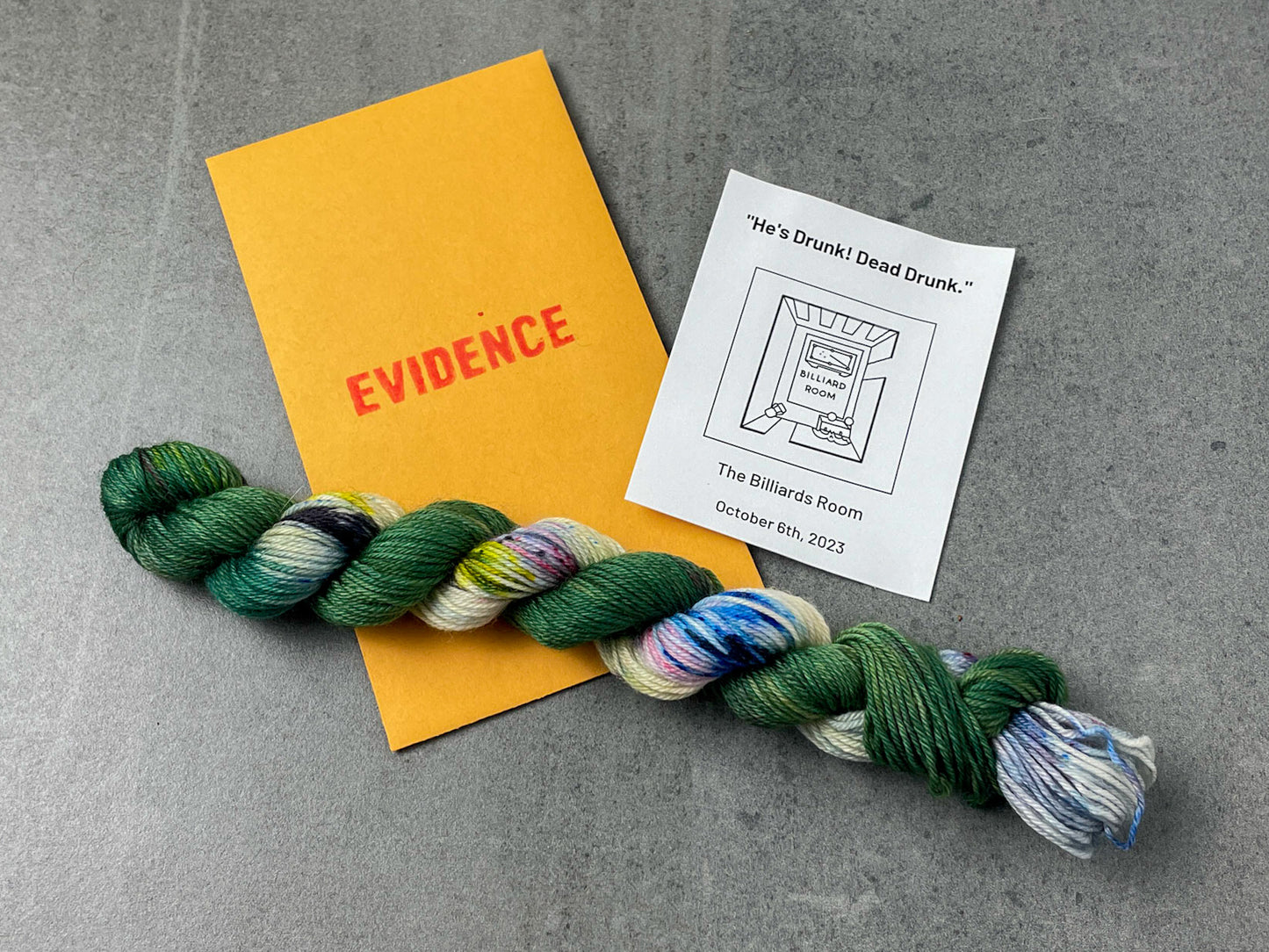 A skein of green and speckled yarn on top of an envelope stamped with "Evidence" and a card with a birds-eye drawing of the billiard room on it.