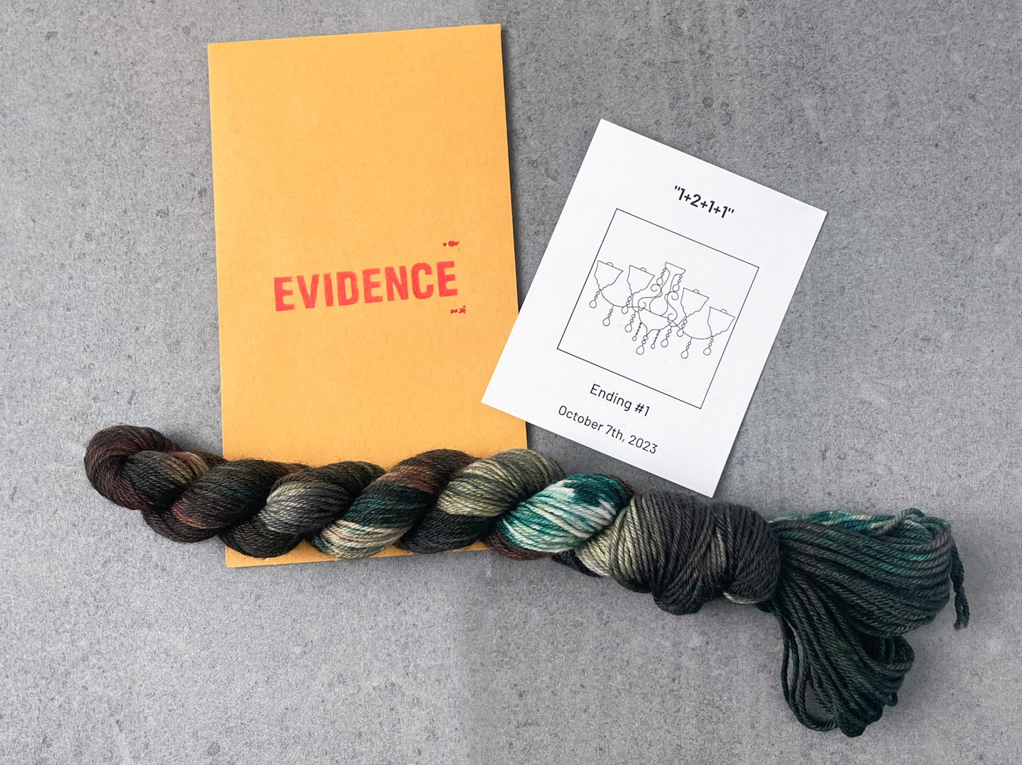 A skein of brown, green, and tan yarn on top of an envelope stamped with "Evidence" and a card with a drawing of a chandelier on it.