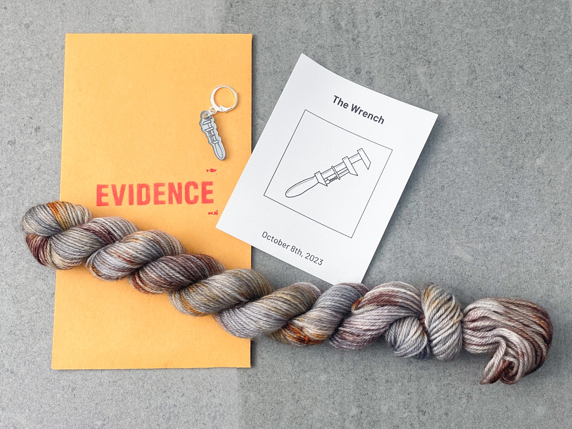 A silver-gray skein with rusty red and brown speckles on top of an envelope stamped with "Evidence" and a card with a drawing of a wrench on it.