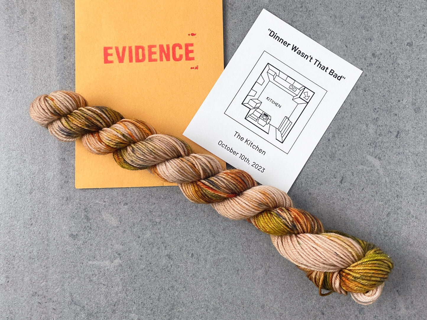 A tan skein of yarn with brown, yellow, orange, and black speckles on top of an envelope stamped with "Evidence" and a card with an overhead drawing of the kitchen on it.