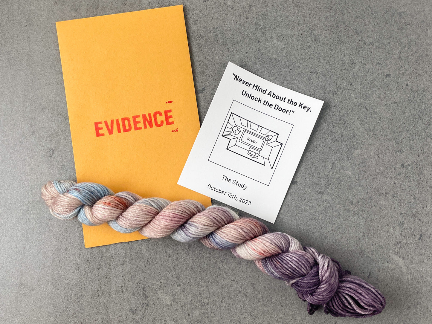 A skein of pastel pink, blue, and purple yarn on top of an envelope stamped with "Evidence" and a card with a drawing of the study on it.
