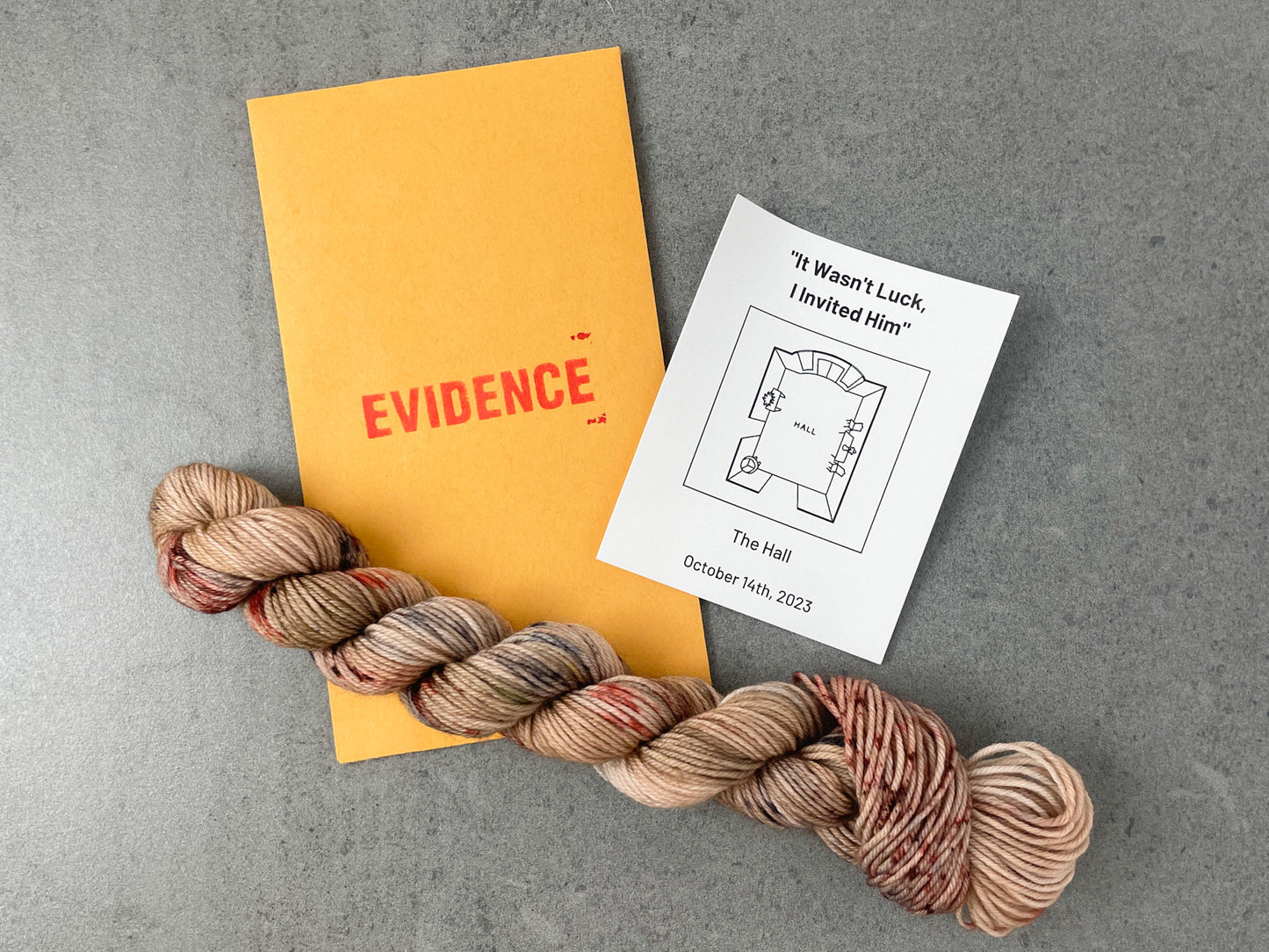 A skein of tan yarn with brown, black, and blood-red speckles on top of an envelope stamped with "Evidence" and a card with an overhead drawing of the hall on it.
