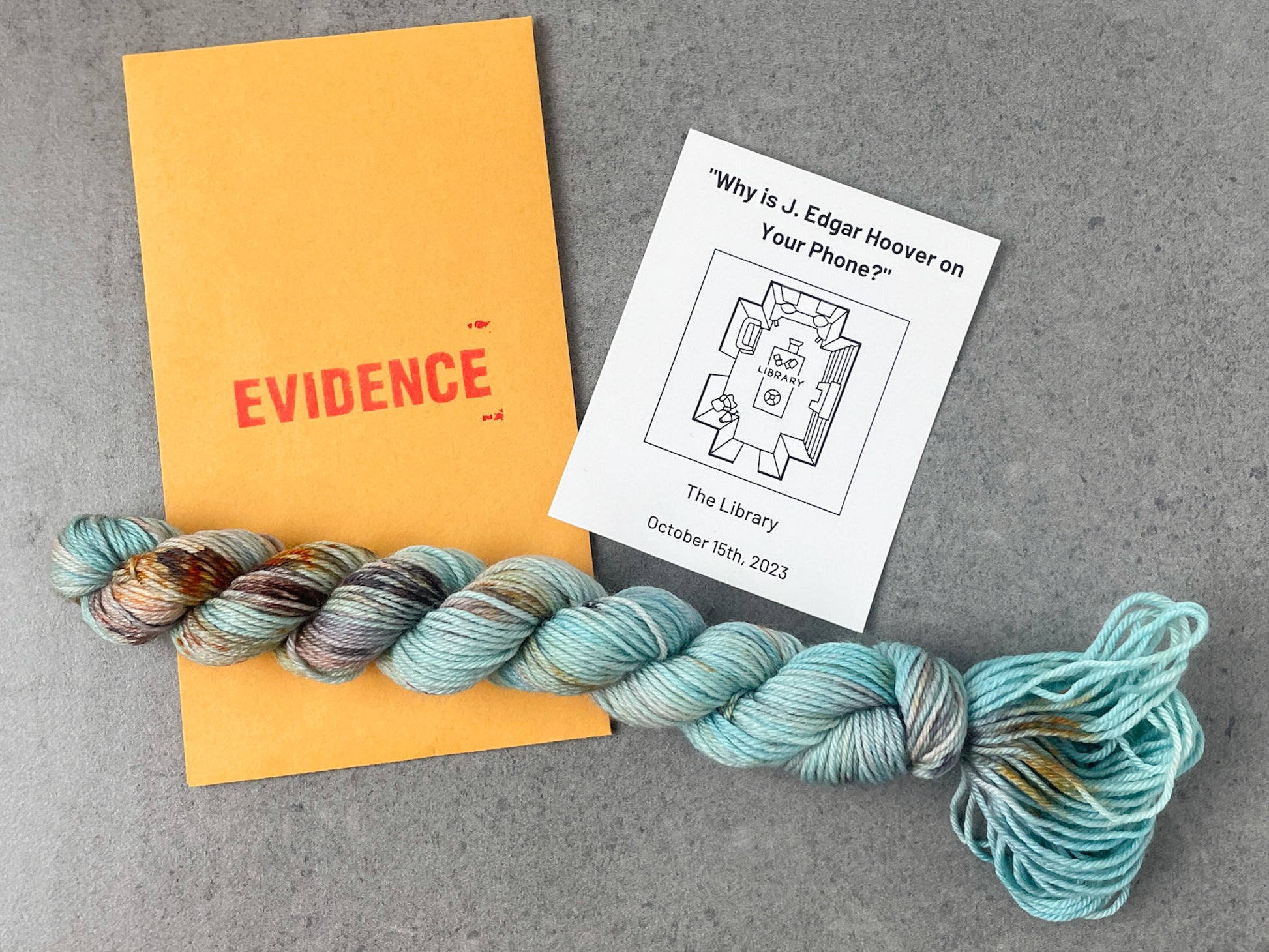 A skein of blue yarn with gray, green, and red speckles on top of an envelope stamped with "Evidence" and a card with an overhead drawing of the library on it.