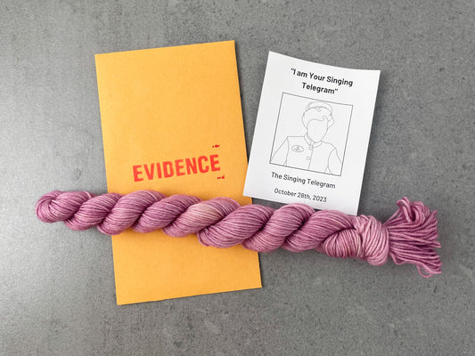 A skein of variegated pink yarn on top of an envelope stamped with "Evidence" and a card with a drawing of the Singing Telegram on it.