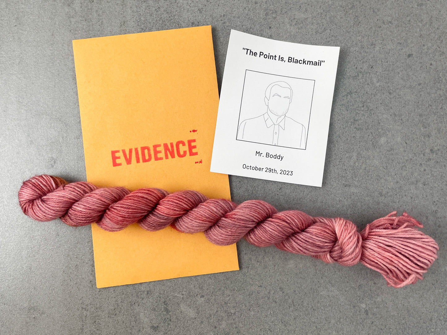 A skein of red-leaning pink tonal yarn on top of an envelope stamped with "Evidence" and a card with a drawing of Mr. Boddy on it.