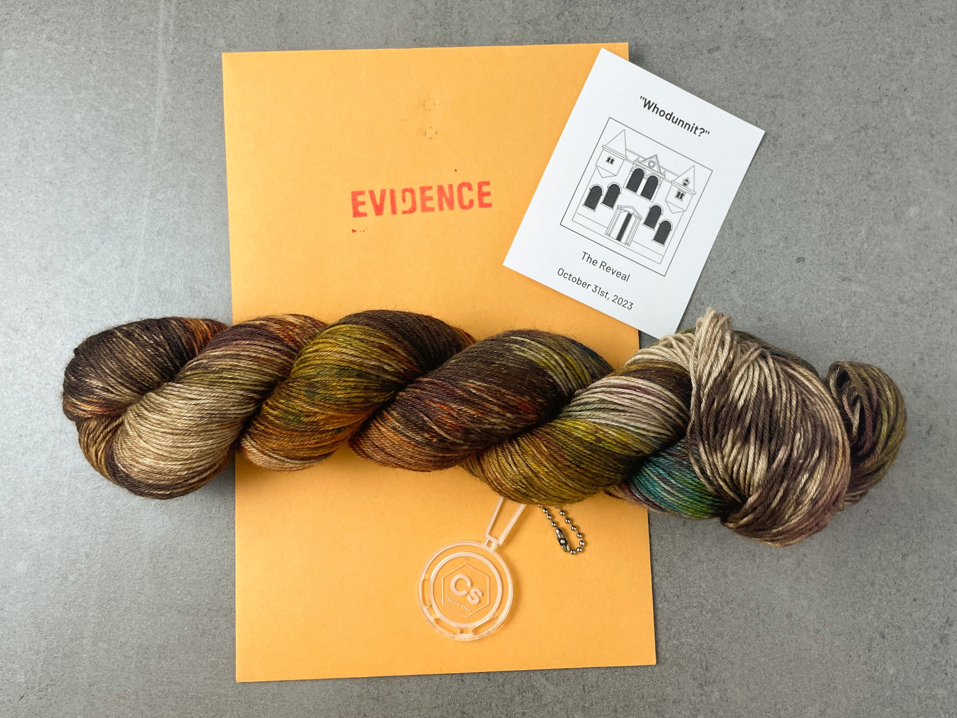 A skein of brown, yellow, tan, and green variegated yarn on top of an envelope stamped with "Evidence" and a card with a drawing of the Clue mansion on it.