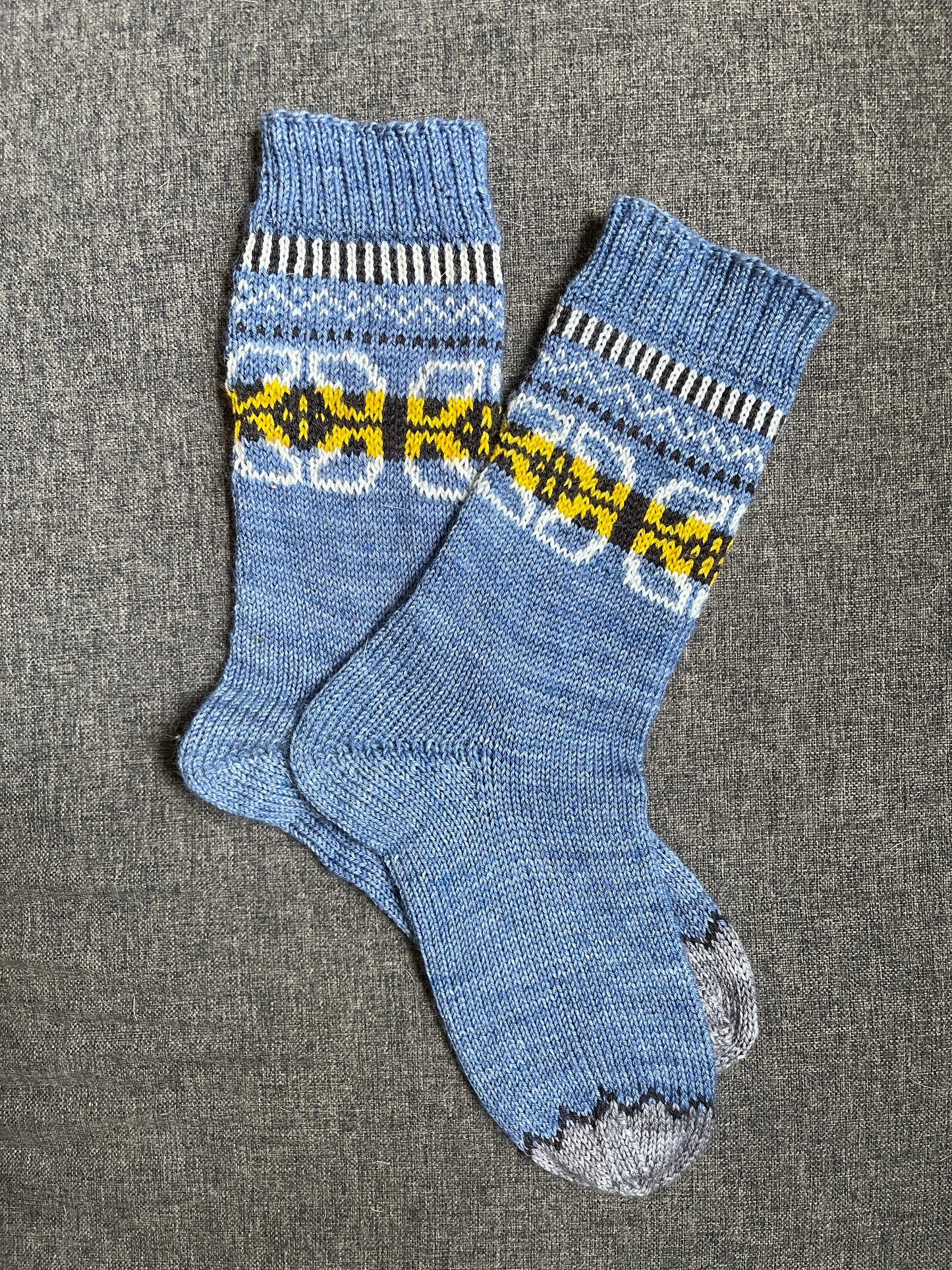 A pair of blue knit socks with a floral pattern, several geometric elements, and a gray toe with a black zigzag border.