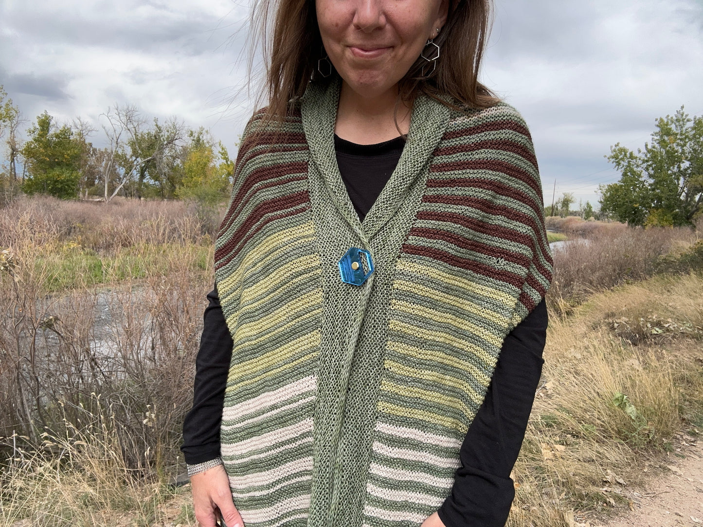 Shana wears the Multistate wrap around her shoulders in a green main color with a blue mirror Shawl Sandwich fastening it at the collar.
