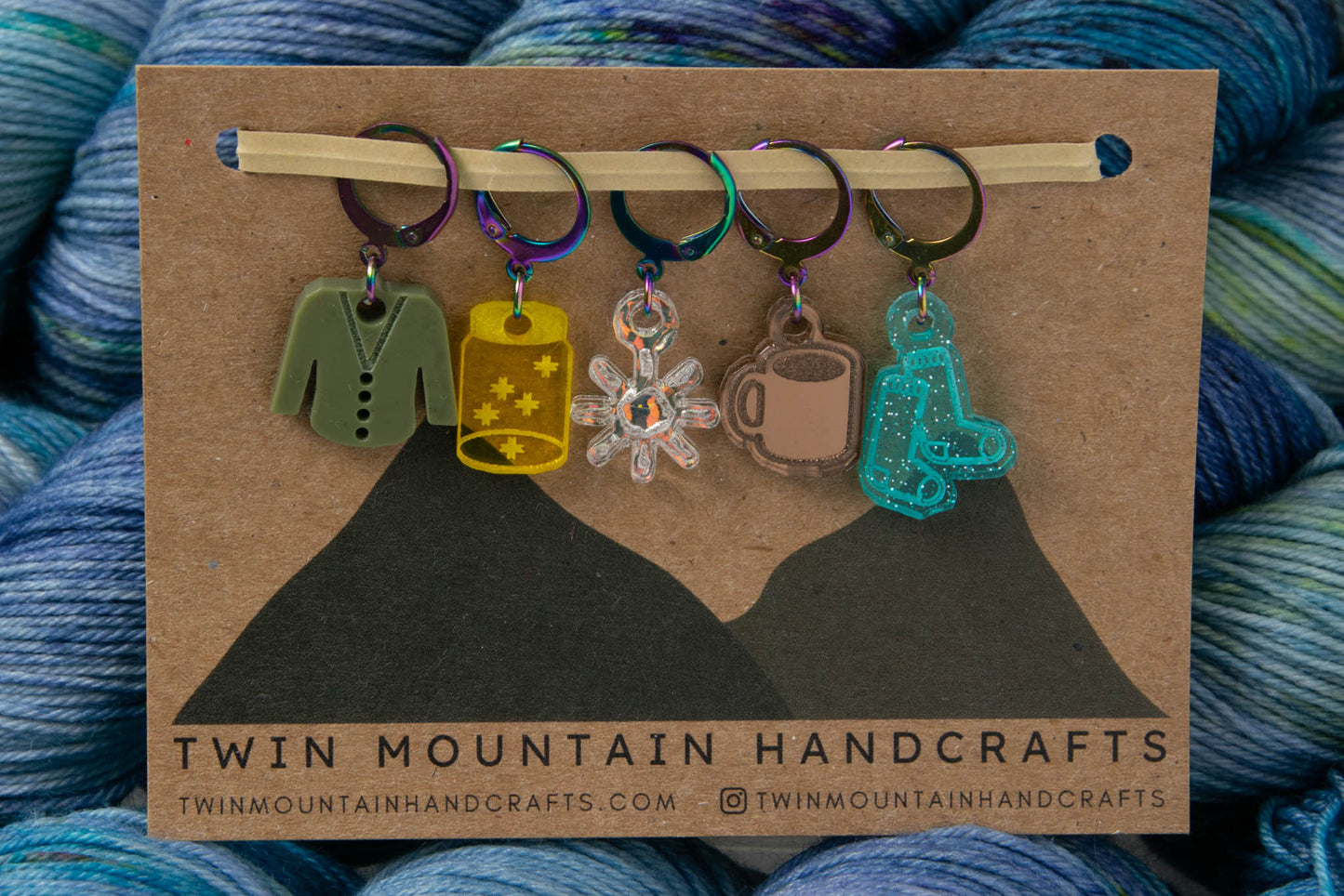 Five stitch markers with rainbow clasps: a green cardigan, a yellow jar with stars in it, an iridescent sun, a tan cup of coffee, and blue sparkly socks.