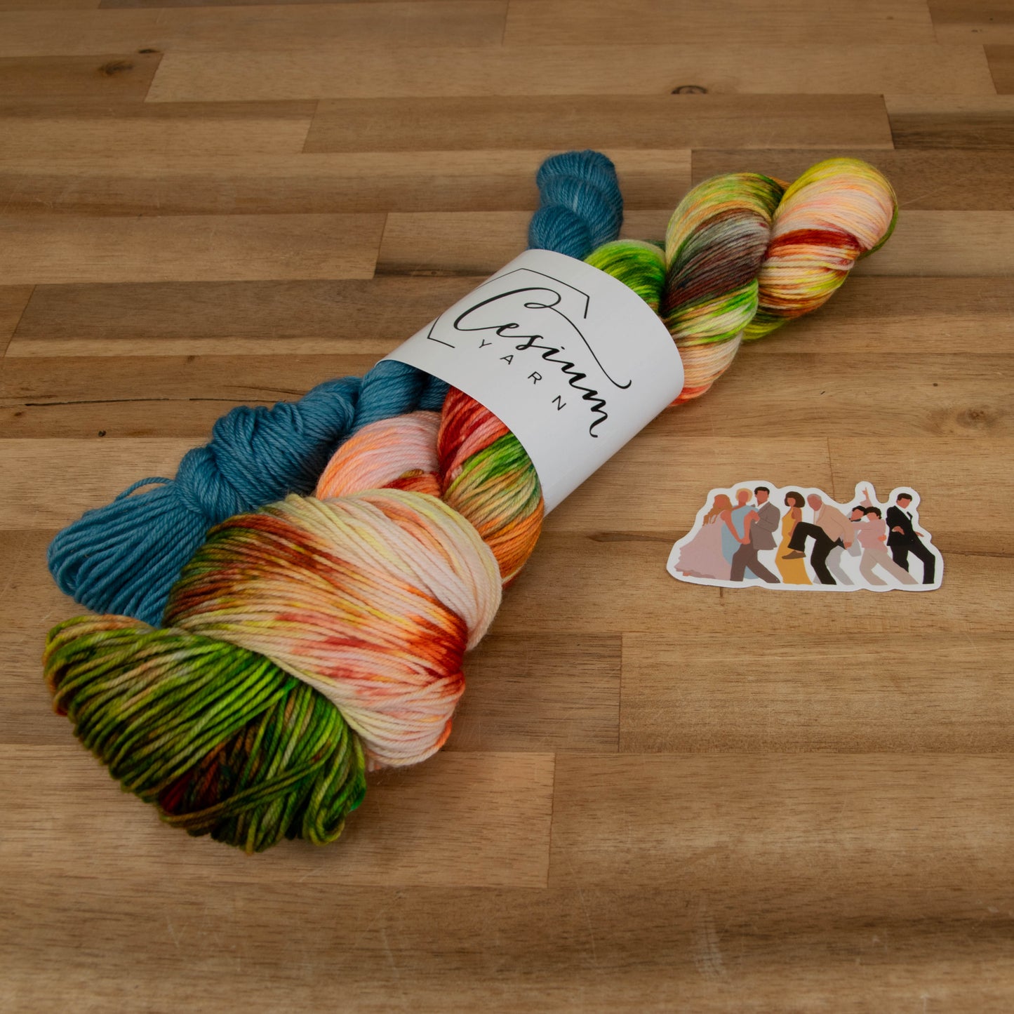 A neon skein of yellow, orange, and green yarn with dark green and red next to an aqua mini skein and a sticker with an illustration of the cast from Mamma Mia.