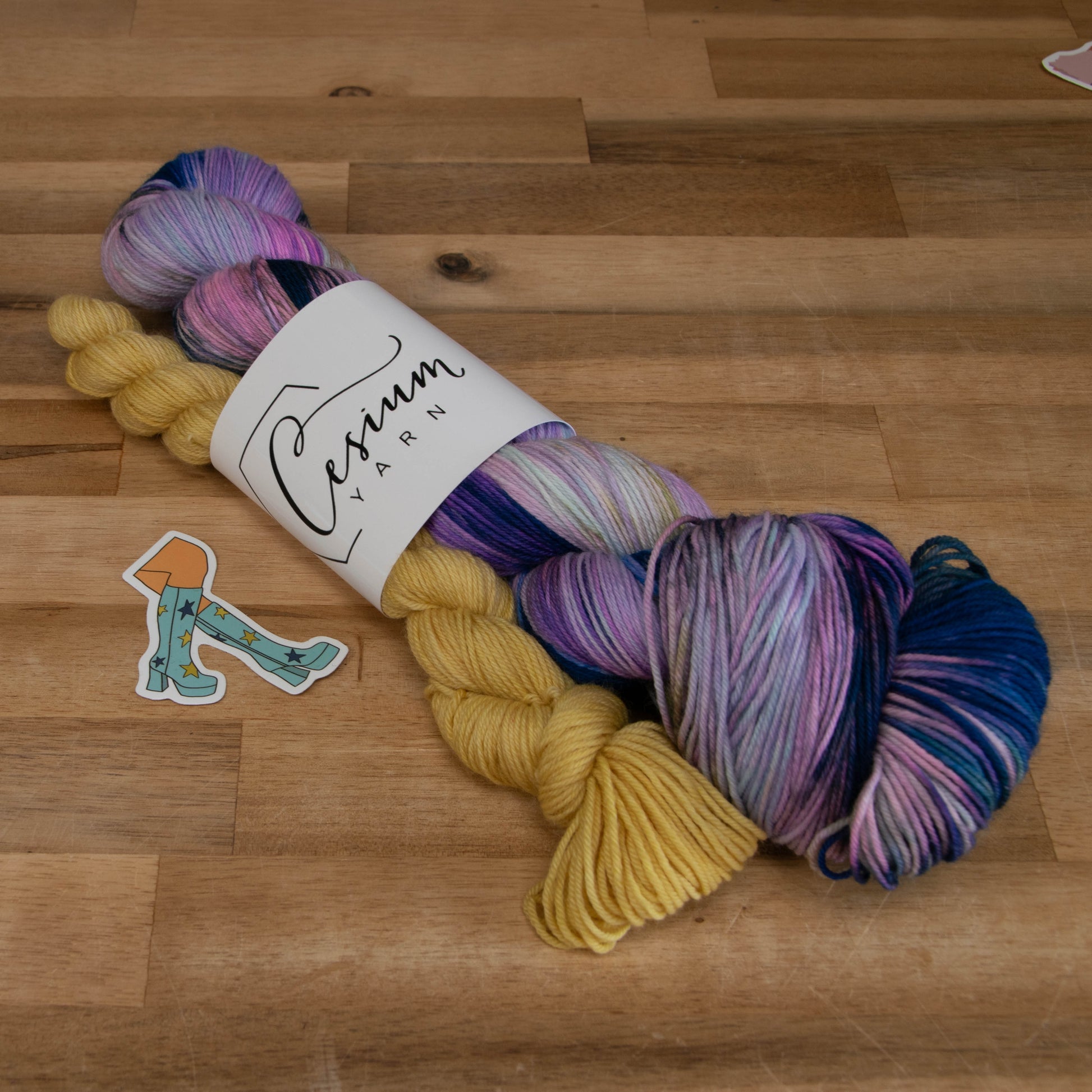 A skein of purple, blue, and pink yarn with a yellow mini skein and a sticker of the Go-Go boots from Mamma Mia.