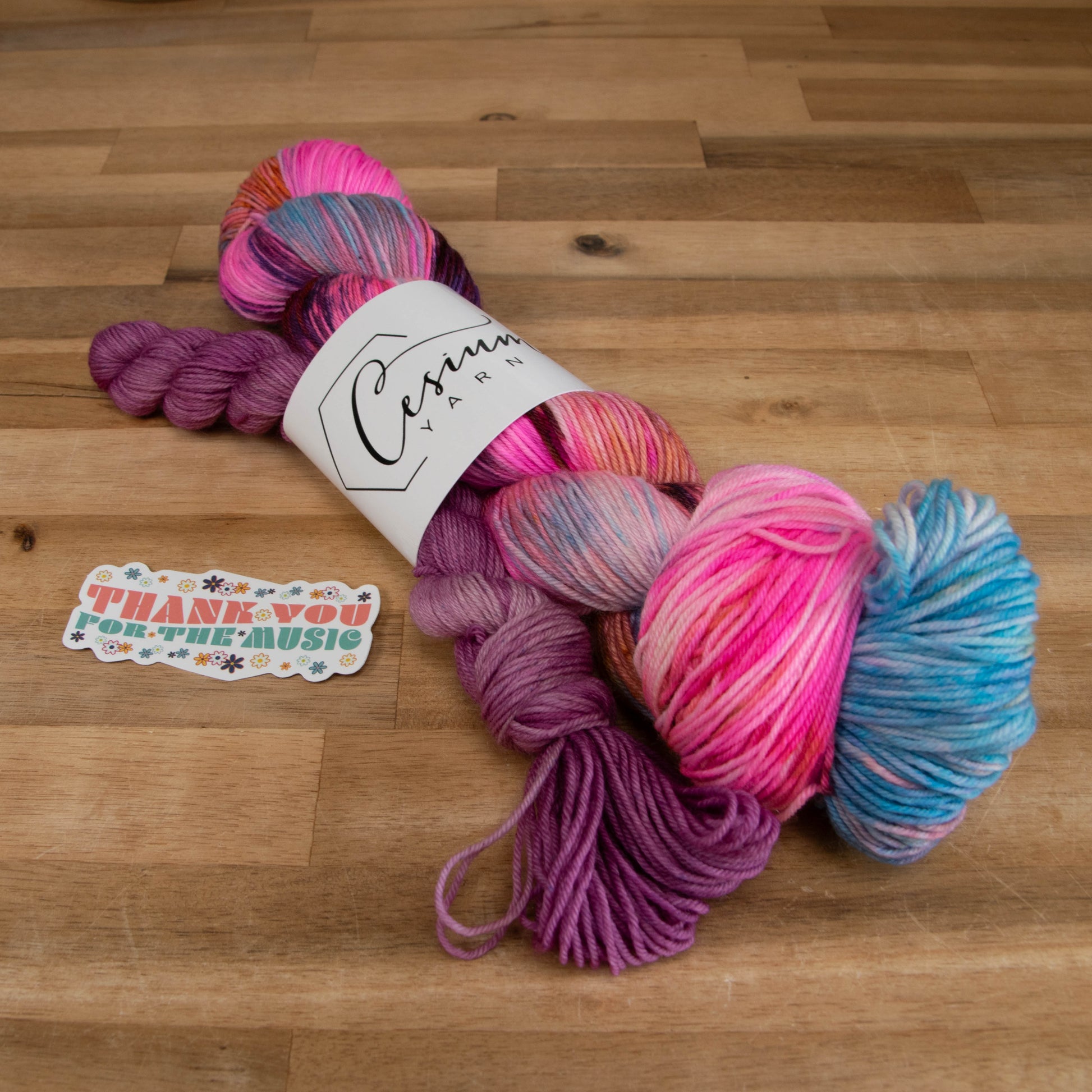 A skein of bright pink, blue, orange, and purple yarn next to a purple mini skein and a sticker that says "Thank you for the music."