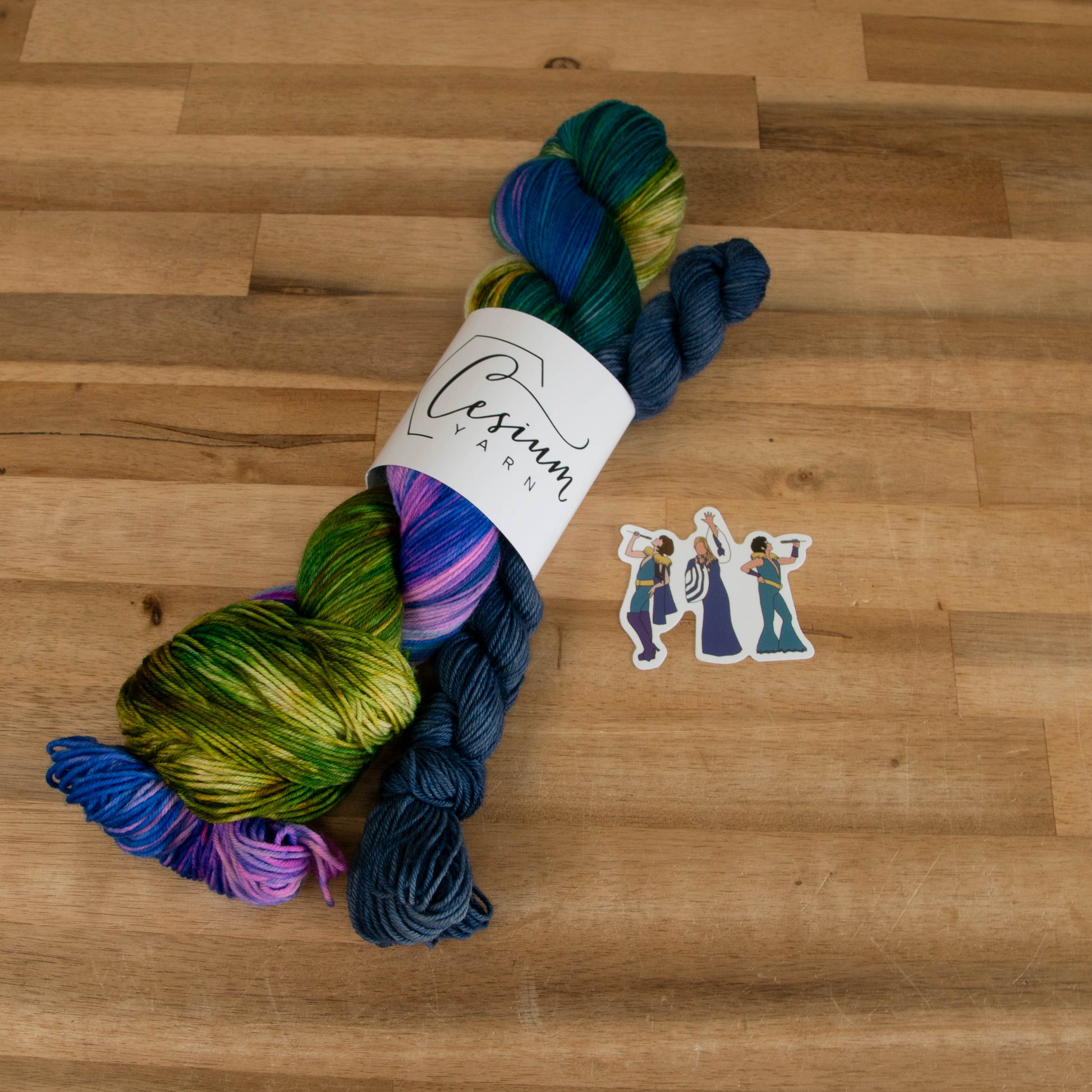A skein of blue, green, and yellow yarn with pink accents next to a denim blue mini skein and a sticker of an illustration of Donna, Tanya, and Rosie.