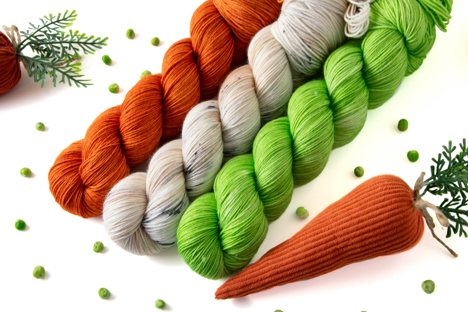 Three skeins of yarn on a white background surrounded by peas and stuffed decorative carrots. The skeins are orange, white with sections of tan and speckled in black, and bright green.