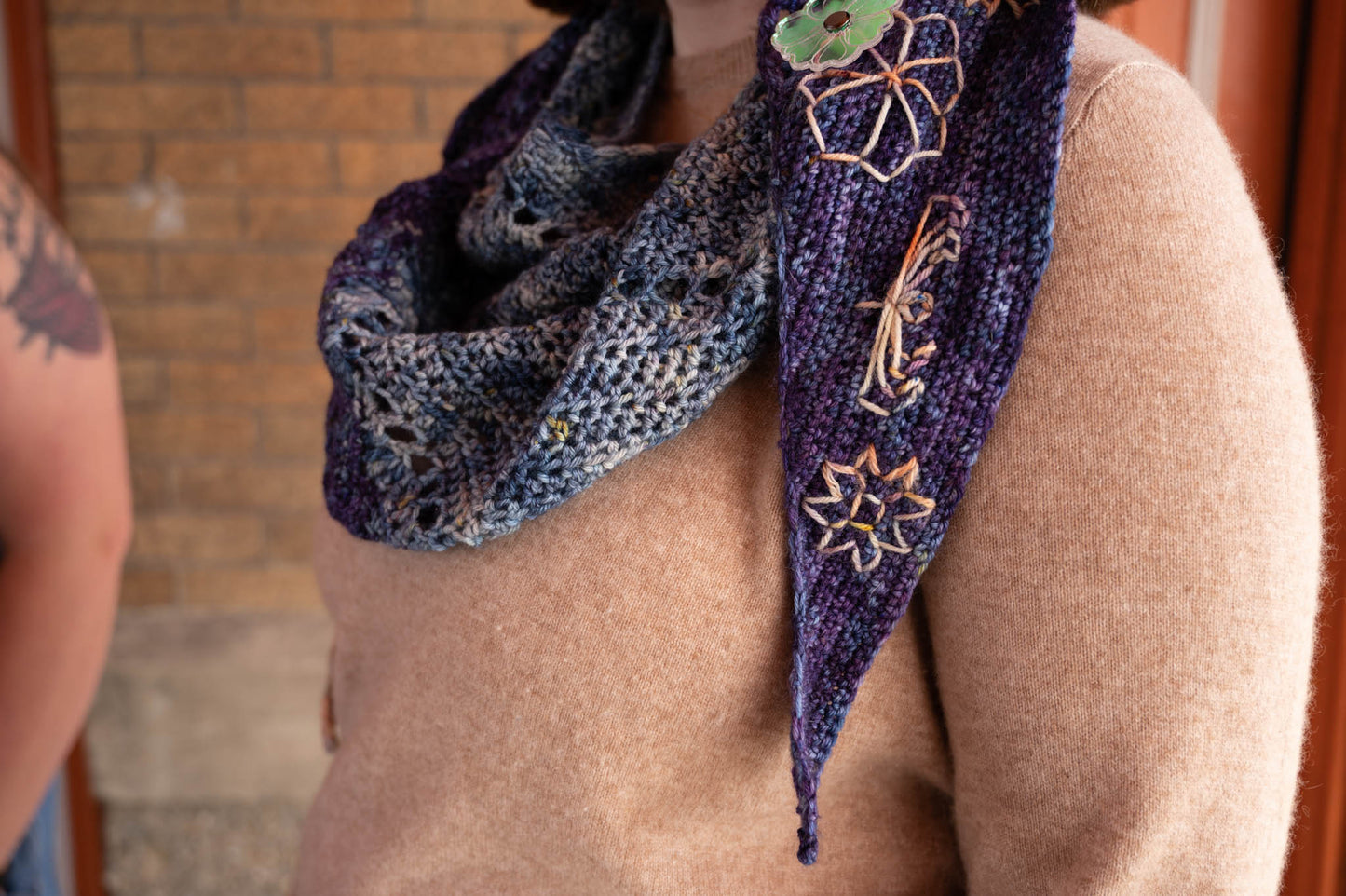 Cat, a white woman, wears a blue and purple crochet shawl wrapped over a tan sweater and fastened with a floral shawl pin.
