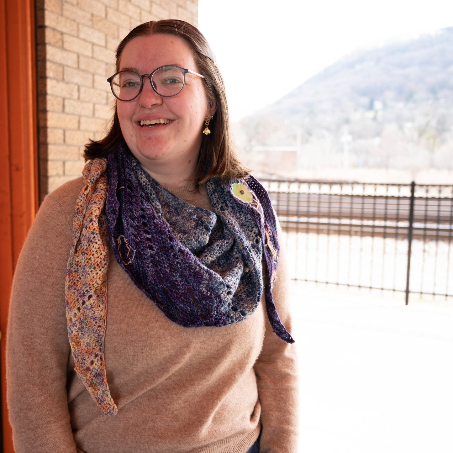 Cat smiles at the camera and shows off the shawl with sections of blue, purple, and a variegated orange, purple, and white yarn.