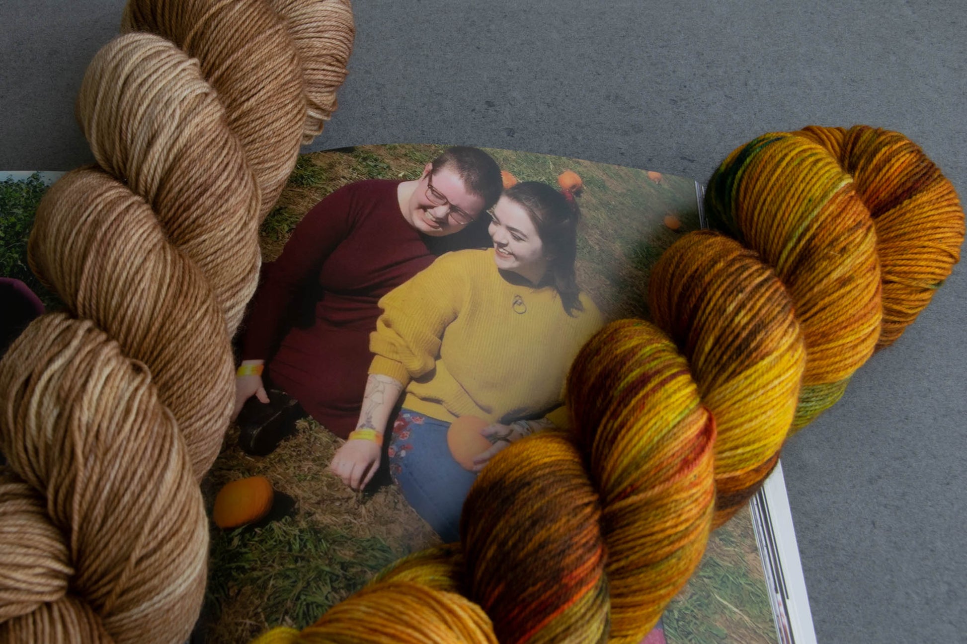 The fall colorways next to a photo of Cat and Penny. An orange, green, and brown variegated skein and one in brown.