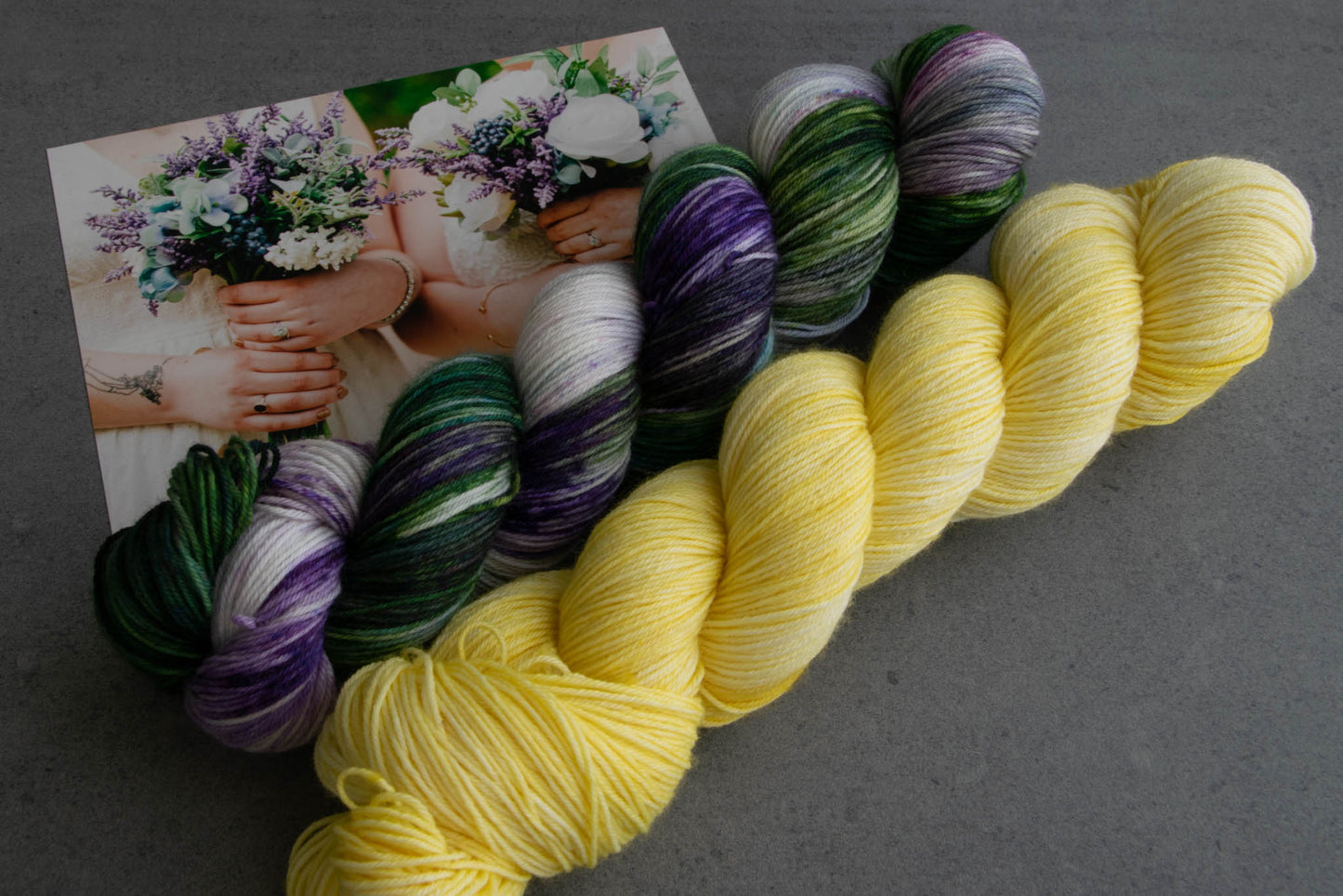 A skein of 4.26 with its complementary purple, green, and white variegated skein and a photo of Cat and Penny holding their wedding bouquets.