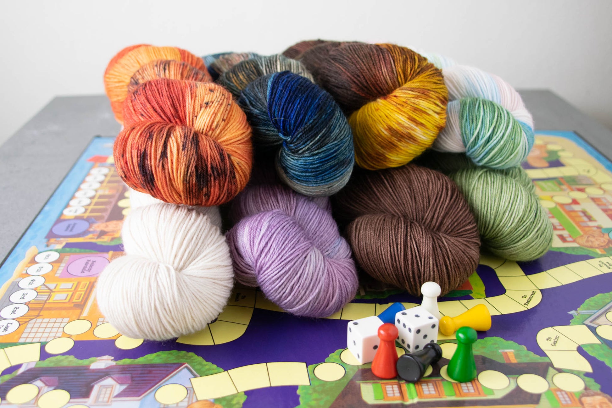 Eight skeins of hand-dyed wool yarn on a vintage board game with wooden pieces and dice.