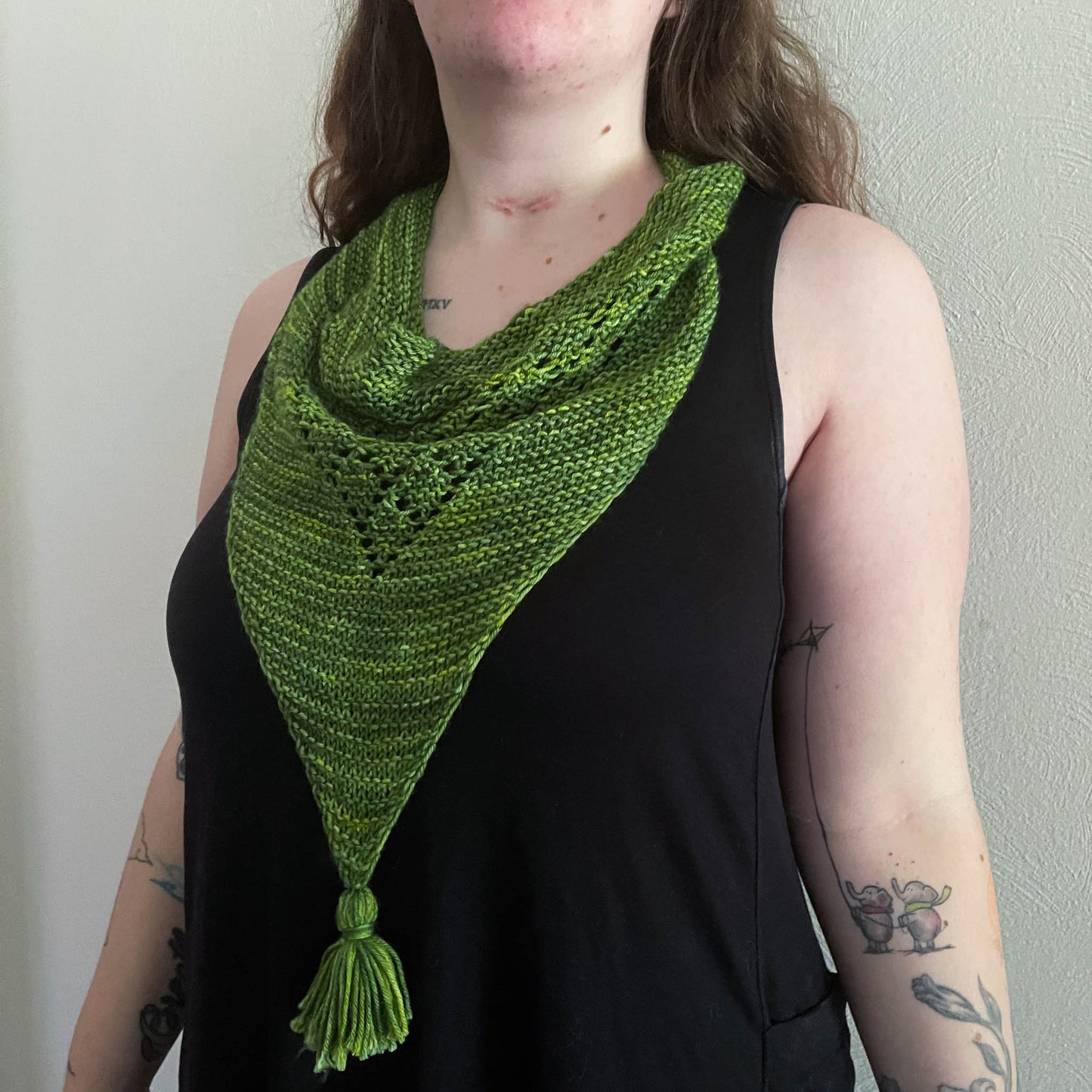 A white woman with tattoos wears a bright green knit bandana cowl with lace details and a pom-pom.
