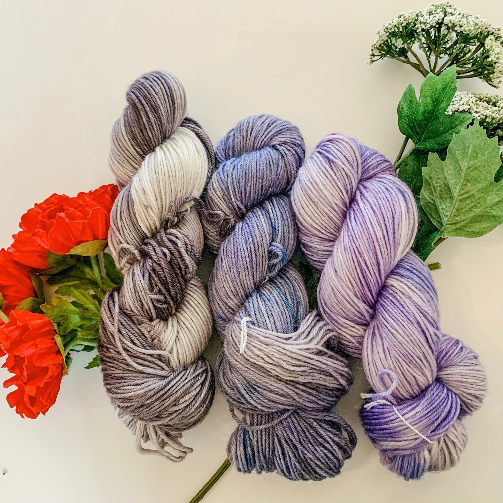 Three cool-toned skeins of yarn from the Poppy collection with poppy and Queen Anne's Lace faux flowers.