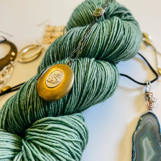 A skein of green hand-dyed yarn with a gold necklace on it and jewelry around it.