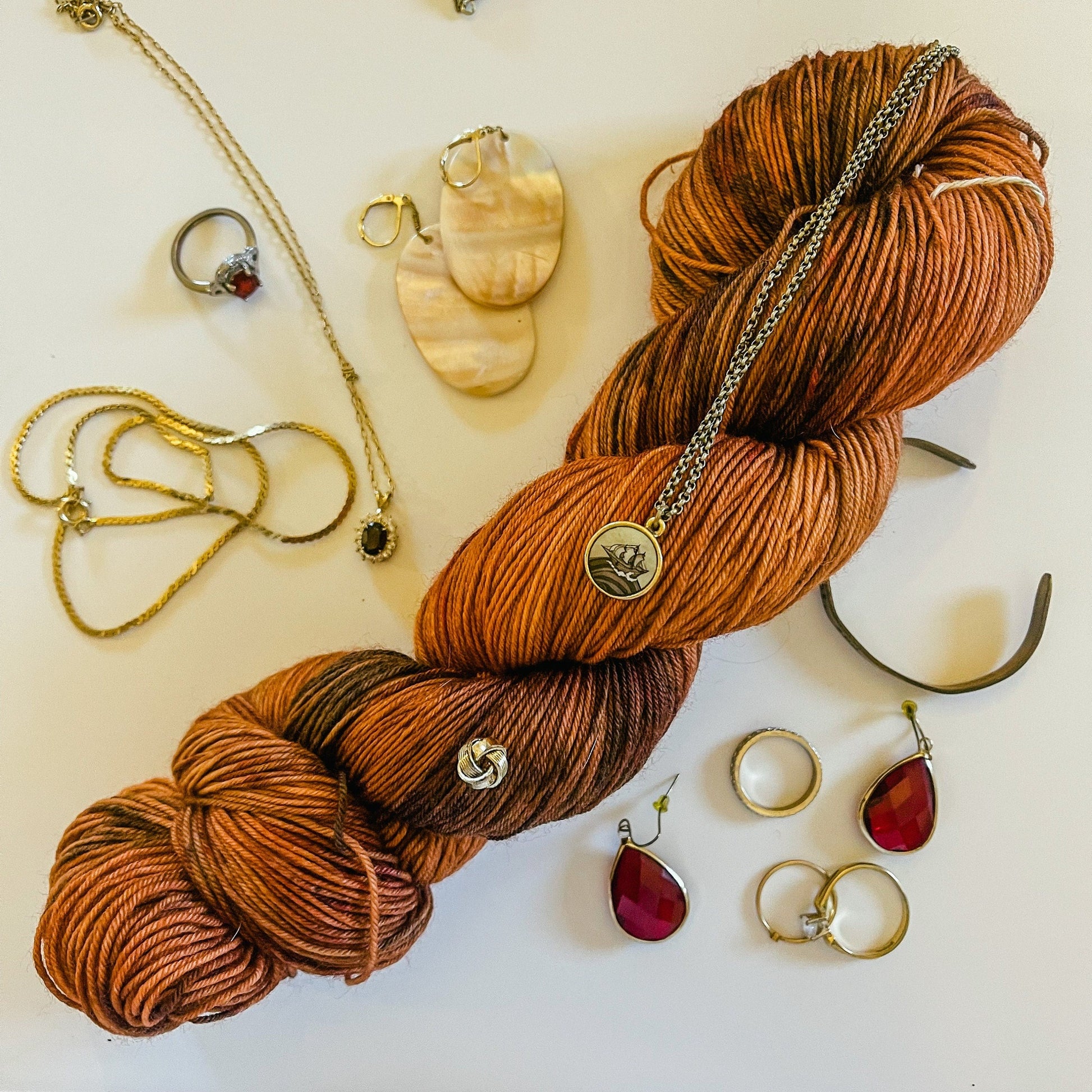 A coppery dark orange skein of  hand-dyed yarn with wide black speckles, surrounded by gold jewelry.