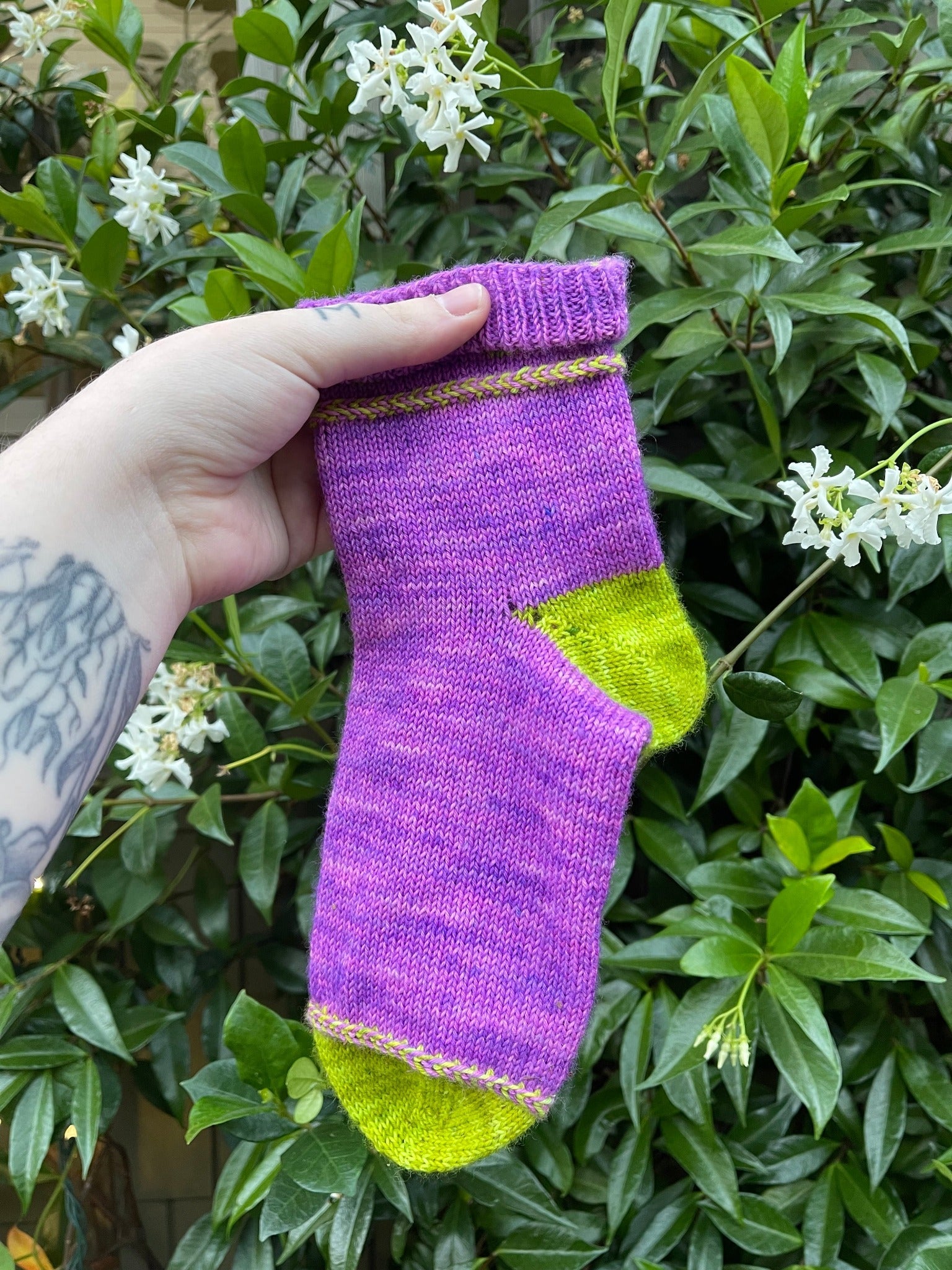 A knit sock with purple as the main color and green as the heel, toe, and braided detail on a background of honeysuckle.